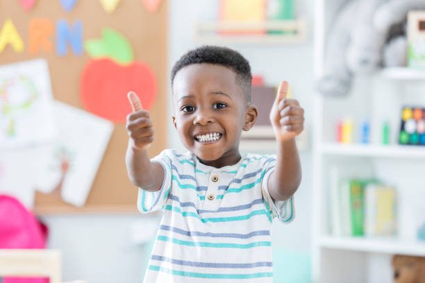 An adorable preschool age little boy smiles for the camera as he stands in his preschool classroom and gives a thumbs up.  He loves school!