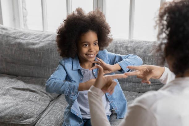 Happy girl talks to disabled deaf mom. Mother teaches kid sign language, shows hand gestures and finger symbols. Tutor giving lesson to positive child with hearing disability at home. Children therapy